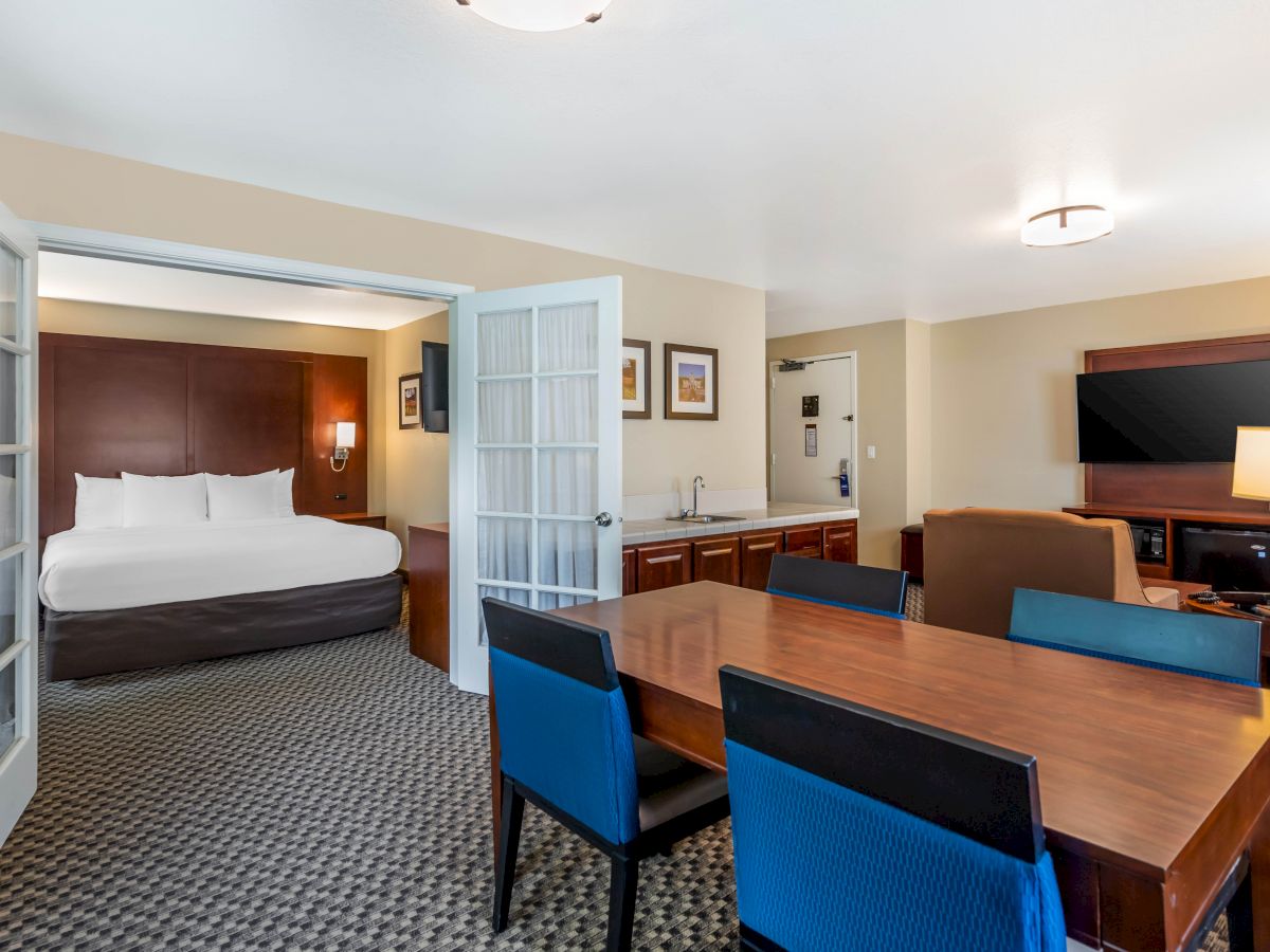 A hotel suite features a bedroom with a large bed, a living area with a TV, a wooden dining table with blue chairs, and a kitchenette area.